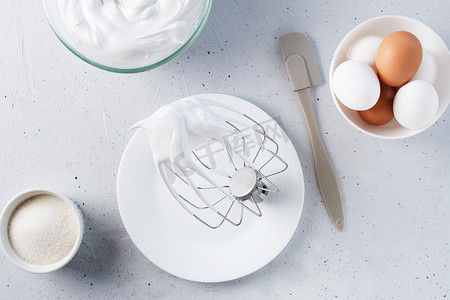 打糖摄影照片_搅打过的蛋清 — 在灰色背景上用金属丝搅拌器、鸡蛋、糖搅打意大利蛋白酥皮。