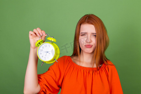 绿色背景中身穿休闲橙色衬衫的年轻红发女性拿着闹钟困倦疲惫