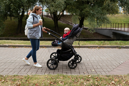 女性咖啡摄影照片_一位 30 岁的母亲推着婴儿车在公园散步，同时喝着杯子里的咖啡