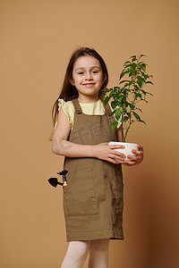 米色背景下拿着花椒植物和园艺工具的白人小女孩梦想成为园丁