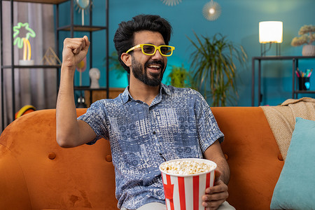 印度男子坐在沙发上吃爆米花，在家看有趣的电视连续剧、在线体育游戏