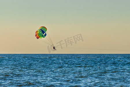 在夕阳的天空和地平线的背景下，男女游客在海上滑翔伞飞行。
