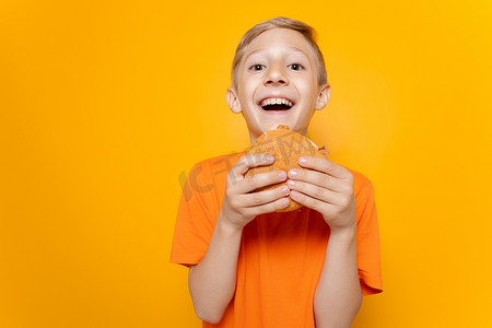 一个身穿橙色 T 恤的男孩在他面前拿着一个汉堡包，在黄色背景下大笑