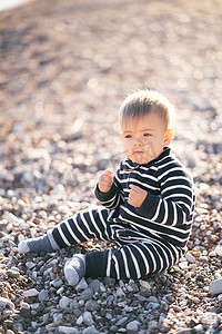 穿着条纹工装裤的严肃小孩坐在鹅卵石滩上，双手握在他面前的拳头上