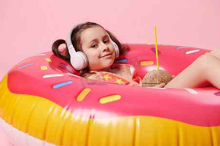 戴着无线耳机的可爱女婴，对着镜头微笑，在泳圈上摆着椰子鸡尾酒，粉红色背景