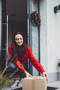 圣诞快递箱摄影照片_身穿红色圣诞毛衣的微笑年轻女子从前门拿起快递箱