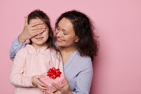 快乐慈爱的母亲闭上可爱女儿的眼睛，同时在孤立的粉红色背景中给她一份可爱的礼物