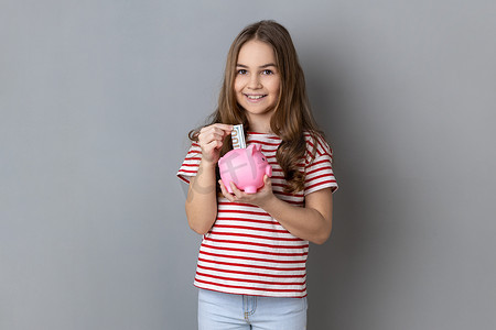 穿着条纹T恤的小女孩站着，手里拿着钞票放进粉红色的存钱罐，