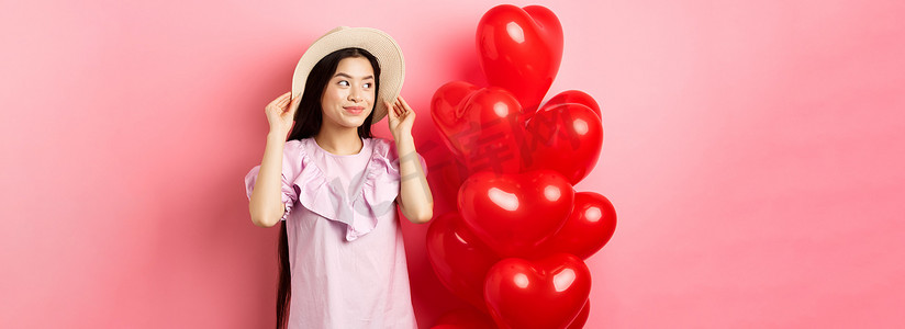 温柔浪漫的亚洲少女约会时戴着草帽和裙子，站在情人节心形气球附近，带着梦幻般的微笑、粉红色背景望向一旁