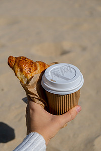 美味的新鲜羊角面包，在海滩大海背景下，女性手中的纸杯中装有咖啡。