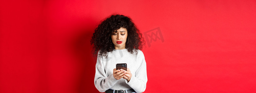 悲伤而忧心忡忡的女人在智能手机上阅读信息，在社交媒体上收到坏消息，站在红色背景下