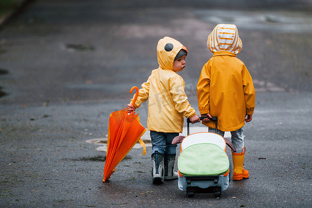 两个带着雨伞、手提箱和黄色防水斗篷和靴子的孩子在雨后一起在户外散步