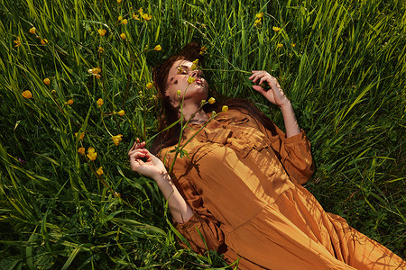 一个长着红色长发的平静女人躺在黄色花朵的绿色田野里，穿着橙色的连衣裙，闭着眼睛，用手抚摸着草叶，享受着平静和休养