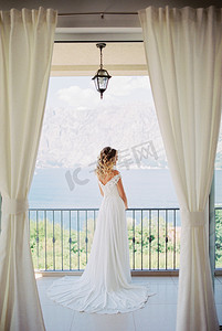 一件白色礼服的新娘在旅馆房间的阳台站立并且看海