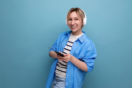 条纹毛衣摄影照片_一位身着条纹毛衣和休闲衬衫、戴着白色无线耳机、面带微笑的金发年轻女子的特写照片在蓝色背景的智能手机中的应用程序中进行通信