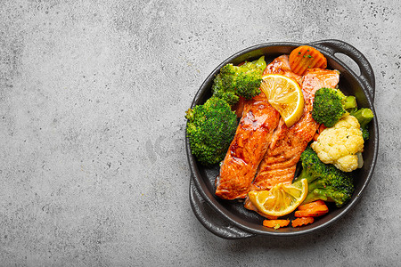 健康的烤鱼鲑鱼排、西兰花、花椰菜、胡萝卜在灰色石头背景的黑色铸铁砂锅碗中。