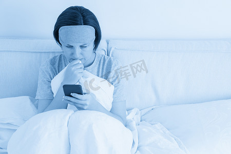 睡眠手机摄影照片_戴着睡眠面具的女人坐在铺着羽绒被的床上，用智能手机发短信