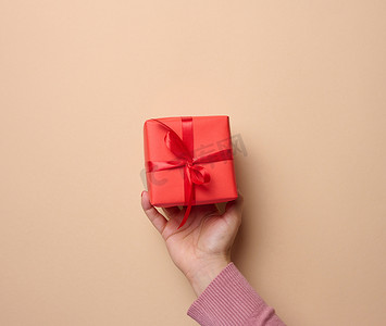 一个女人的手拿着一个红色礼盒，上面裹着米色背景的丝带，这是一个祝贺、惊喜的概念