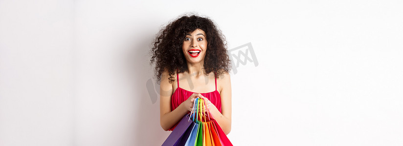 促销活动黑金摄影照片_穿着时尚红裙、拿着购物袋微笑、在促销活动中购买、站在白色背景上的快乐年轻女性