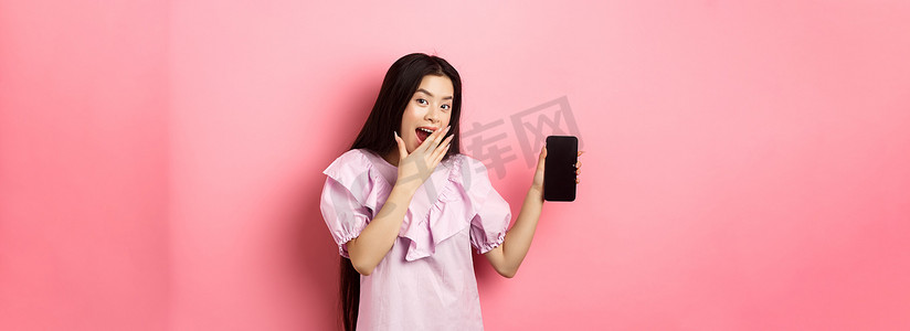 欢快的亚洲少女展示着空荡荡的手机屏幕，笑着用手捂住嘴，穿着粉色背景的裙子