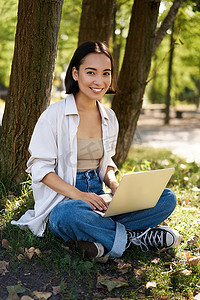 美丽的亚洲女孩带着笔记本电脑坐在公园里，在遥控器上工作，在键盘上打字，对着镜头微笑，在树旁休息
