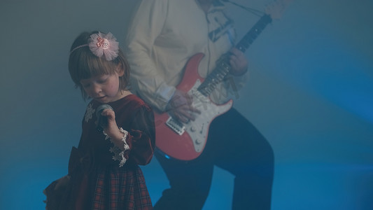 重汽开业舞台图摄影照片_穿着复古裙子的小女孩在舞台上唱歌，她的父亲弹着电吉他