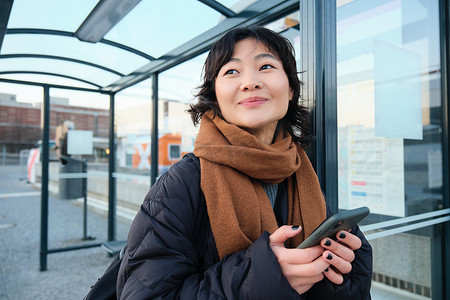 漂亮的日本女孩，学生穿着冬装站在公交车站，拿着智能手机，使用应用程序跟踪她的公共交通，叫车