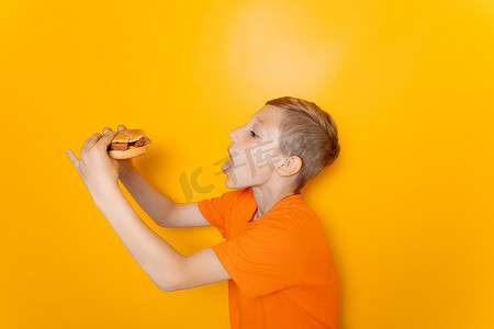 一个穿橙色T恤的男孩侧身站着，手里拿着一个他想吃的汉堡包