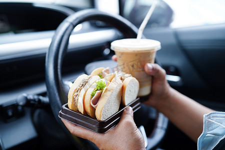 亚洲女司机拿着冷水和三明治面包在车里吃喝，很危险，有发生事故的风险。