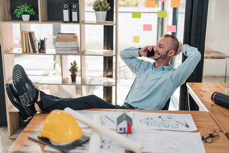英俊而快乐的白人男性工程师将双腿放在桌子上，手放在脑后，同时与客户通电话，在办公室里放松。