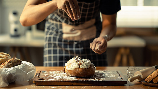 男面包师穿着围裙在面团上筛面粉，在厨房里做面包