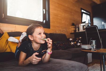 相亲节目摄影照片_快乐的小女孩在电视上看她最喜欢的节目时吃零食