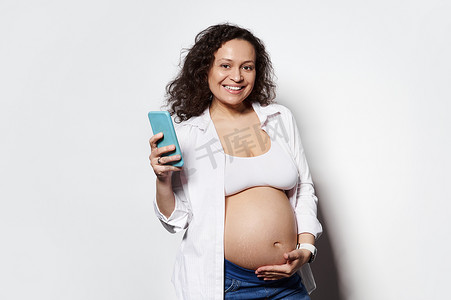 快乐的中年孕妇对着镜头微笑，抚摸着她的肚子，拿着手机，在白色背景中被隔离