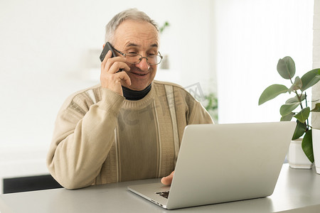 英俊微笑的老人戴着眼镜使用手机，坐在他舒适的工作场所，家里有笔记本电脑，退休男性在社交媒体上与朋友聊天，在智能手机上打字。