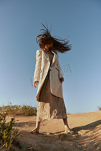 一位身穿时尚夹克和长裙的女士站在沙滩上，背景是晴朗的天空，她的头发在运动中遮住了脸
