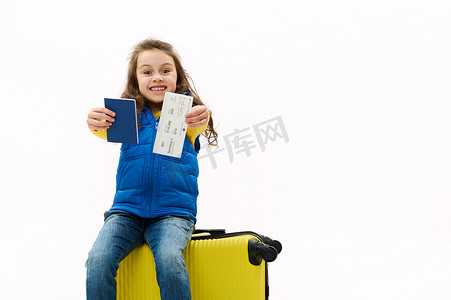 淘气的小女孩坐在手提箱上，拿着护照和机票，为即将到来的出国旅行而高兴