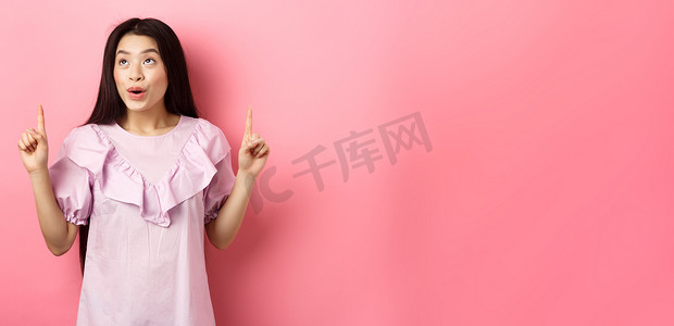 兴奋的亚洲少女竖起手指，看着空荡荡的空间，穿着粉红色背景的裙子，看起来很梦幻
