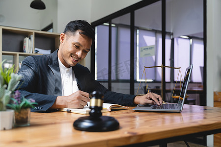 身着正装的职业亚洲商人、律师或财务顾问在办公桌前工作，使用笔记本电脑并在书上做笔记