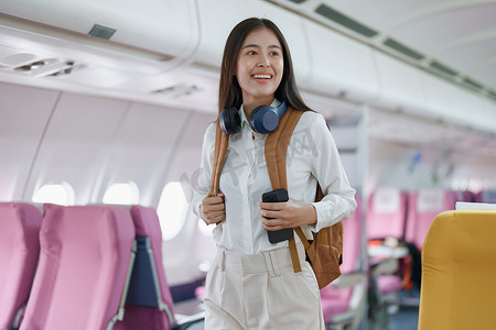年轻的亚洲美女乘飞机旅行，乘客戴着耳机把手提行李放在飞机座位上方的储物柜里