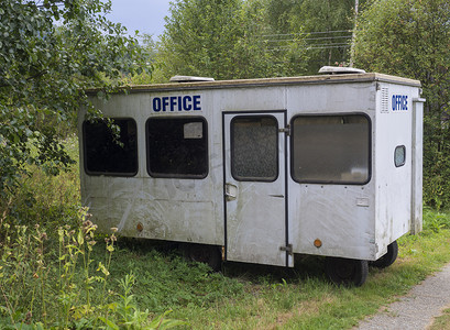 拖车的标志摄影照片_带蓝色标志办公室的白色小拖车房，在靠近人行道、绿草和树木的乡村景观中废弃的大篷车