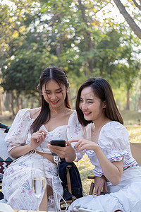 穿着漂亮裙子的轻松亚洲女性正在手机上一起看视频，她们正在野餐，她们谈得很开心