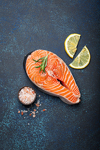 深蓝色质朴石背景上带迷迭香的新鲜生鲑鱼片牛排，玫瑰盐和柠檬顶视图，健康均衡的饮食和营养