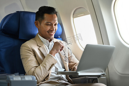 豪华飞机摄影照片_商务旅行期间，穿着优雅豪华西装的成功商人在飞机上使用笔记本电脑