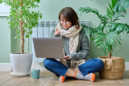 视频秋天摄影照片_暖气片附近穿着暖和衣服的中年女性使用笔记本电脑进行视频通话