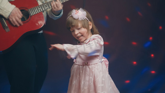 儿童节音乐摄影照片_穿着复古连衣裙的小女孩在舞台上跳舞，她的父亲弹着原声吉他