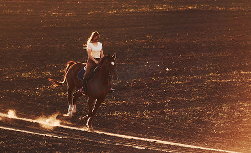 女子骑马摄影照片_阳光明媚的白天，戴着防护帽的年轻女子在农田里骑马