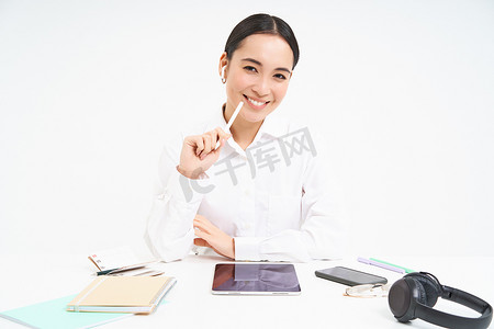 办公室里年轻的亚洲女售货员在数字平板电脑上工作，桌上放着笔记本和耳机，背景是白色