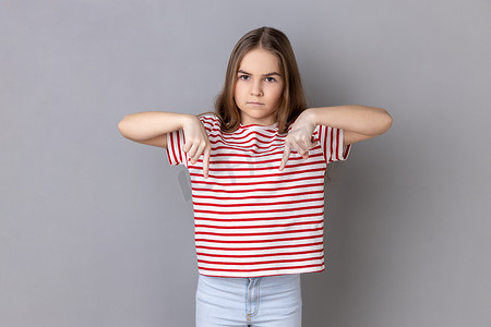 专横的摄影照片_专横的黑发小女孩穿着条纹 T 恤站着，食指向下指着。