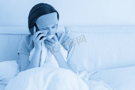 悲伤的女人戴着睡眠面具，坐在床上，用智能手机盖着羽绒被