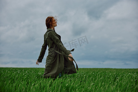 一位身穿长雨衣的红发女子在多云的天气中站在绿地里，在强风中